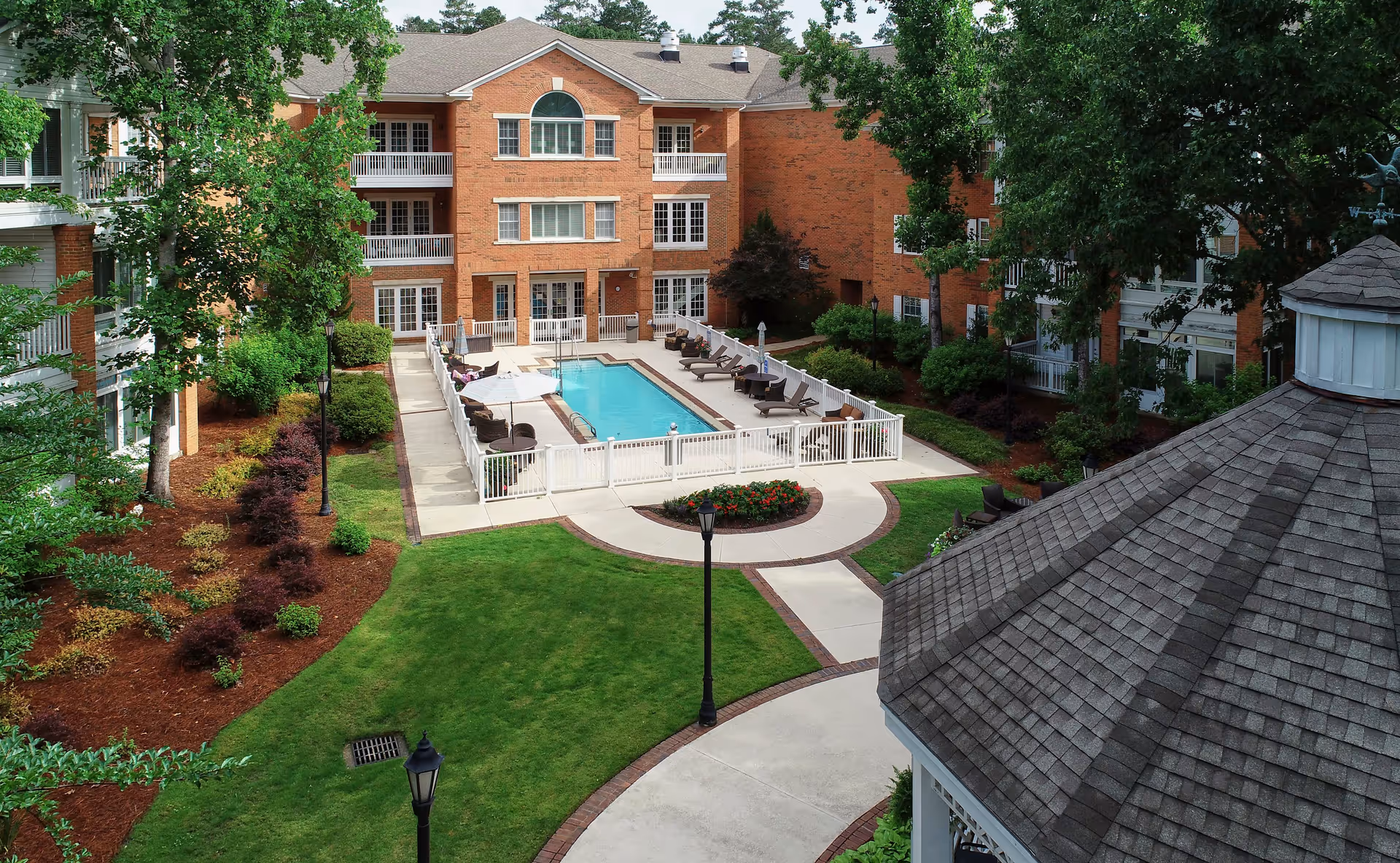 Outdoor courtyard area of a senior living facility named Galleria Woods featuring a rectangular swimming pool surrounded by lounge chairs and umbrellas. The pool area is fenced and centrally located within a landscaped garden with green grass, trees, shrubs, and paved walkways. Surrounding the courtyard are multi-story brick buildings with balconies and windows. A gazebo roof is visible in the foreground.