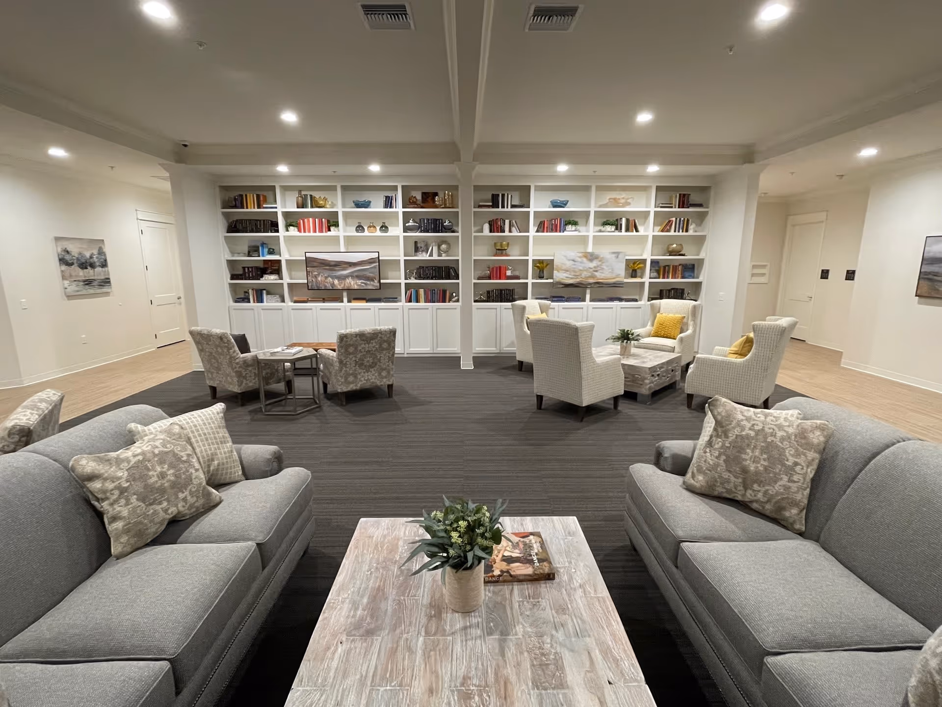 Spacious furnished common living room with sofas, armchairs, coffee tables, and built-in bookshelves along the back wall.