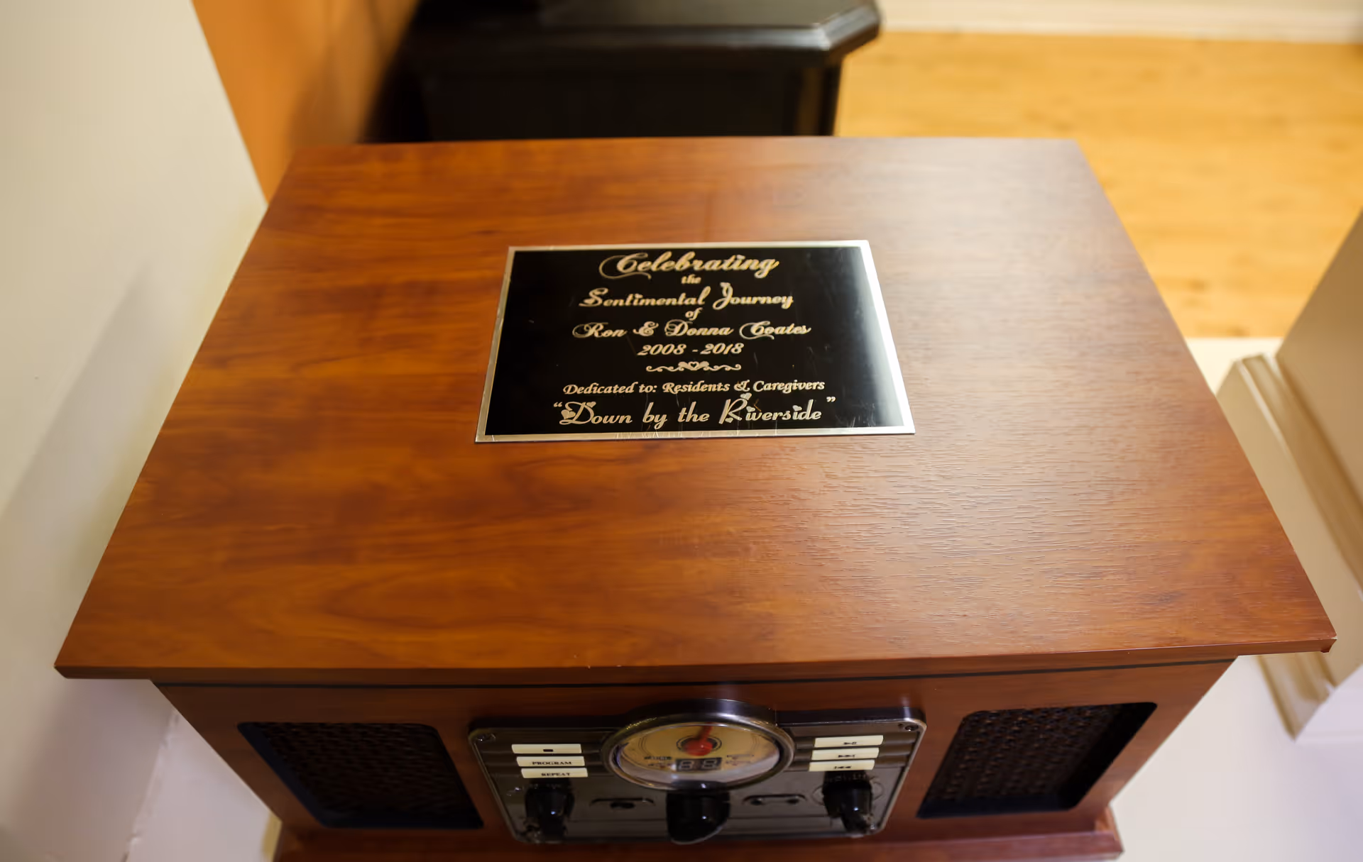 A wooden piece of furniture with a plaque on top that reads: Celebrating the Sentimental Journey Ron & Donna Coates 2008-2018 Dedicated to: Residents & Caregivers 'Down by the Riverside'. The furniture appears to be a vintage-style radio or record player.