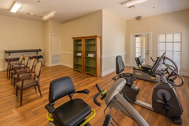 Small fitness room with recumbent exercise bikes, a row of chairs, wooden shelving and hardwood floors.