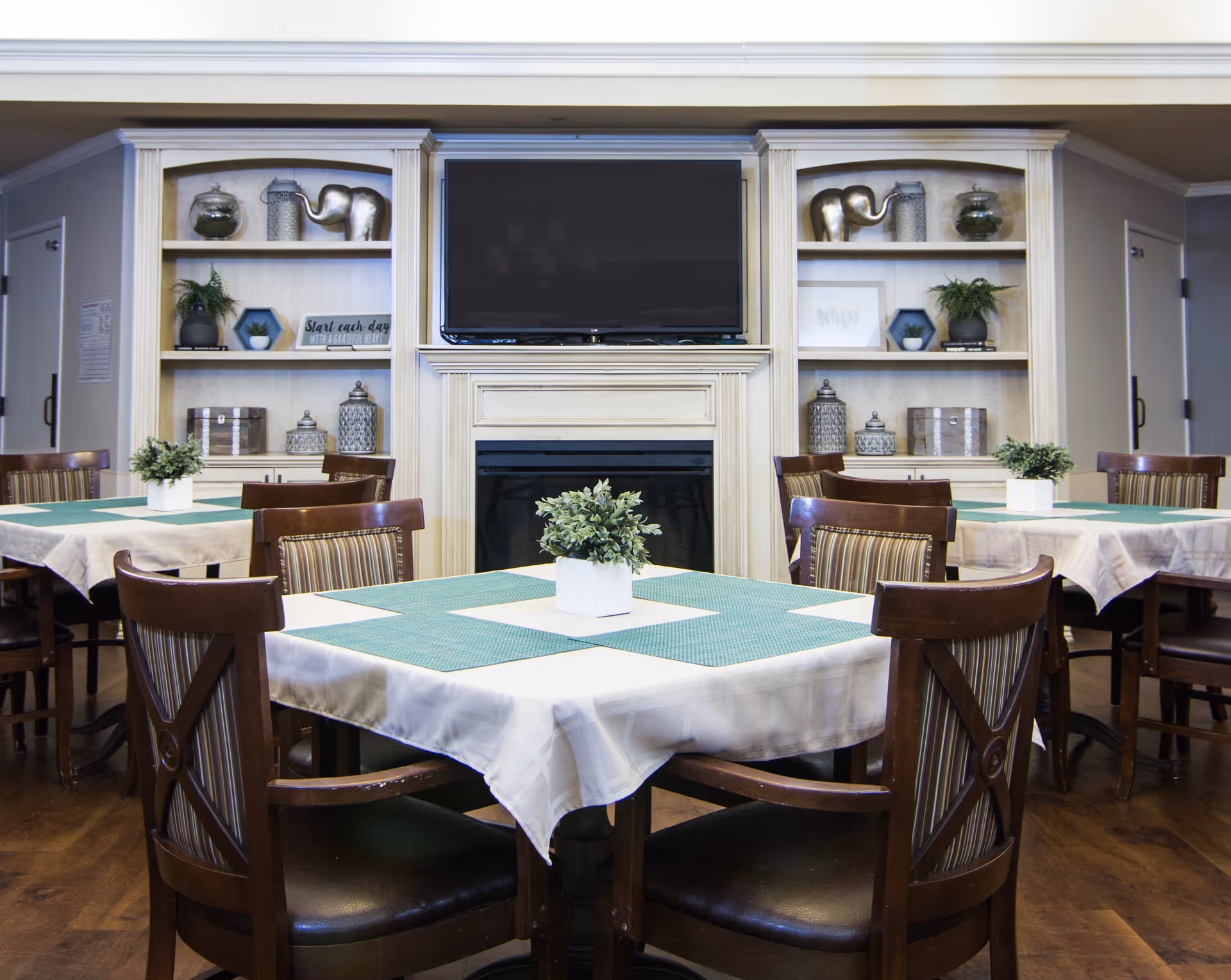 A dining area with several tables covered with white tablecloths and green placemats, each table having a small potted plant centerpiece. The room features wooden chairs with striped cushions, built-in shelves with decorative items, a fireplace, and a large flat-screen TV mounted above the fireplace.