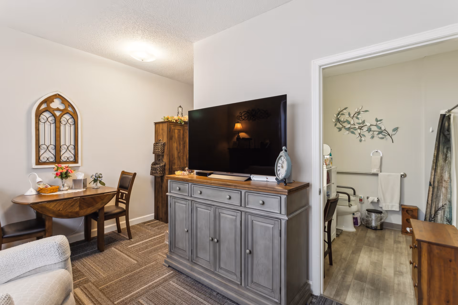 Interior view of a senior living facility room showing a small dining area with a wooden table and two chairs, a gray cabinet with a large flat-screen TV on top, and an open doorway leading to a bathroom with a toilet, grab bars, towel racks, and decorative wall art.
