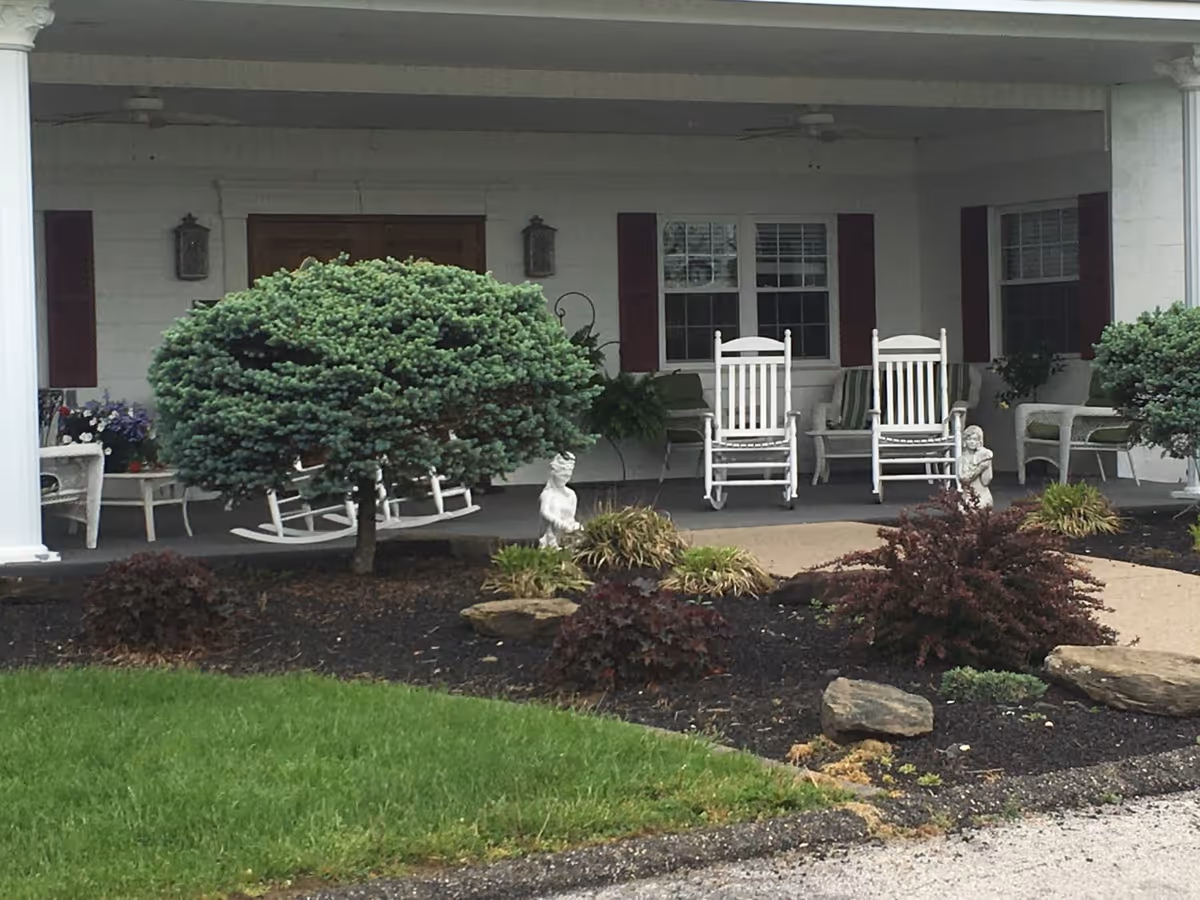 Front porch of a building with white rocking chairs, a small table, and decorative plants and shrubs in the garden bed in front. The porch has white columns and windows with dark shutters.