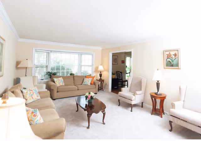 A bright and cozy living room with beige walls and carpet. The room features two beige sofas with colorful floral cushions, two beige armchairs, a wooden coffee table with a flower arrangement, and two side tables each with a lamp. A large window with white blinds lets in natural light, and a framed floral painting hangs on the wall.