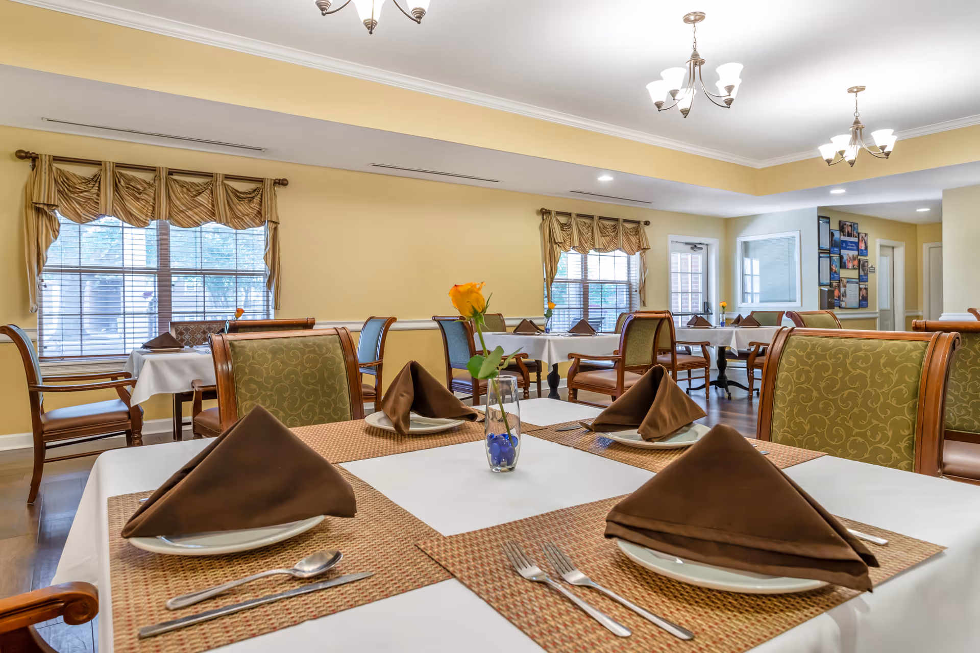 A bright dining room with several tables set for a meal. Each table has white tablecloths, brown napkins folded in a triangle shape, plates, silverware, and a small vase with a yellow flower. The room has large windows with beige curtains, wooden chairs with green patterned cushions, and warm yellow walls. Ceiling lights illuminate the space.