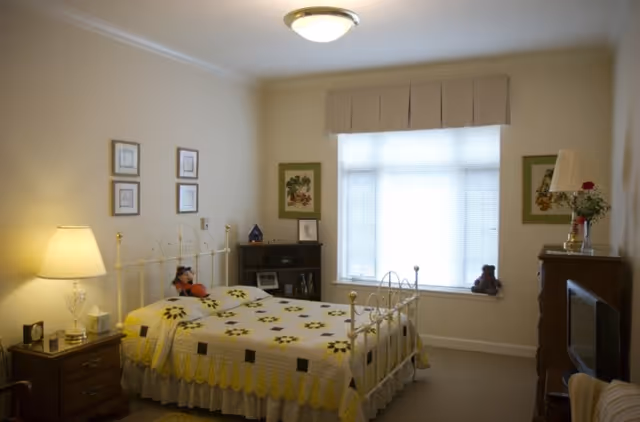 A cozy bedroom with a metal-framed bed covered in a yellow and white quilt. There is a bedside table with a lamp and clock, a small bookshelf, framed pictures on the walls, a window with blinds and a valance, and a dresser with a TV and a lamp with flowers.