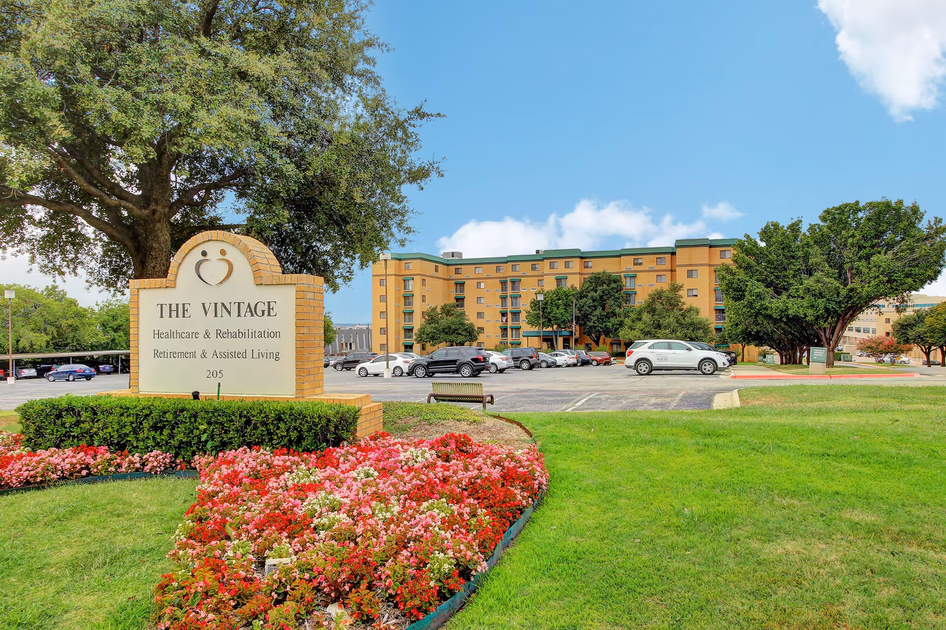 Outdoor view of The Vintage Healthcare & Rehabilitation Retirement & Assisted Living facility with a large sign in the foreground surrounded by colorful flowers and green grass, a parking lot with cars, and a multi-story building in the background under a blue sky with some clouds.