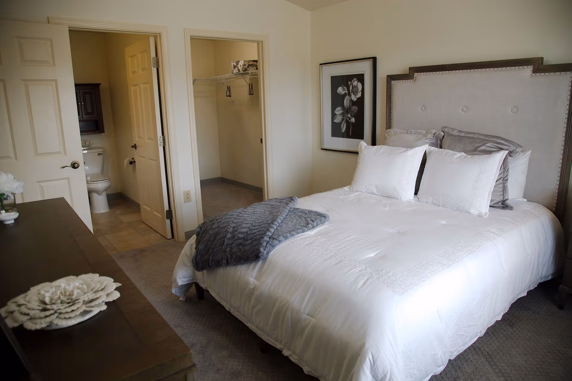 A neatly made bed with white bedding and multiple pillows in a bedroom. To the left, there is a dresser with decorative items on top. Two open doors reveal a bathroom with a toilet and a walk-in closet. A framed black and white floral picture hangs on the wall beside the bed.