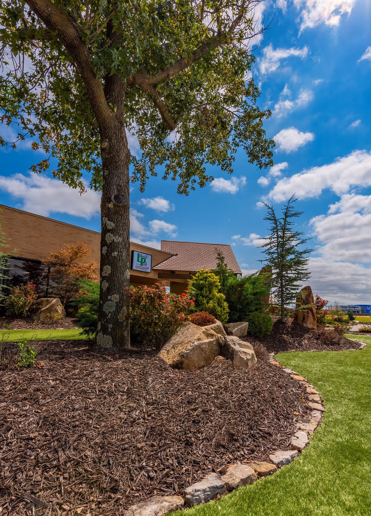 Outdoor landscaped area with a large tree, rocks, shrubs, and a well-maintained lawn in front of a brick building under a partly cloudy blue sky. A sign on the building reads 'Liberty Park Assisted Living Entrance'.