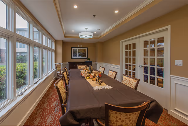 A dining room with a long rectangular table covered with a dark tablecloth and decorated with a centerpiece of flowers and pine cones. The room has multiple upholstered chairs around the table, large windows on one side letting in natural light, beige walls with white wainscoting, a patterned carpet, and a ceiling light fixture. There is a set of French doors on one wall and a framed picture on the far wall.