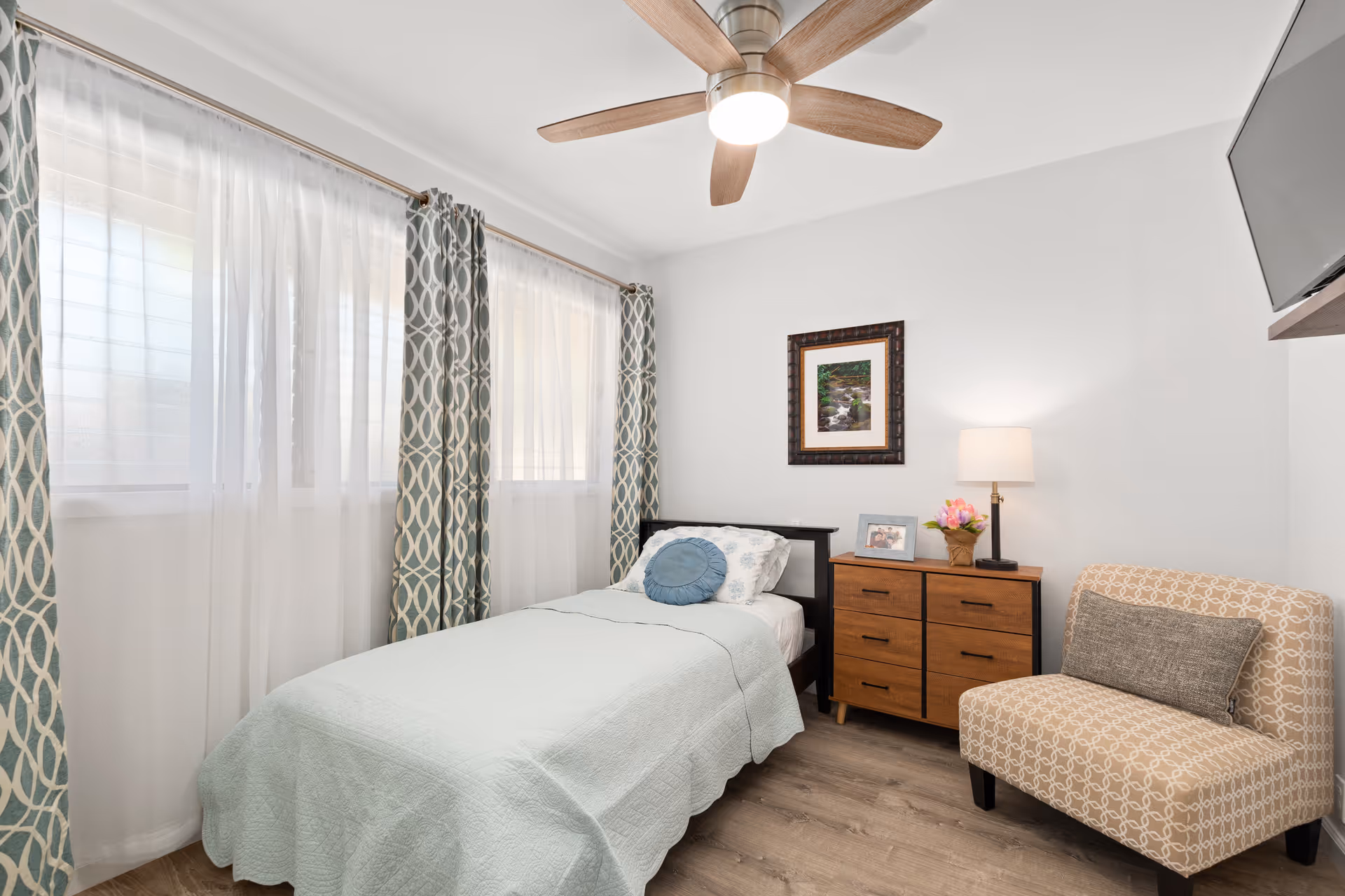 A bright bedroom with a single bed covered in a light blue quilt and a round blue pillow. The room features two large windows with sheer white curtains and patterned drapes. There is a wooden dresser with a lamp, a framed photo, and a flower arrangement on top. A beige patterned armchair with a gray cushion is positioned next to the dresser. A ceiling fan with wooden blades and a mounted flat-screen TV are also visible.