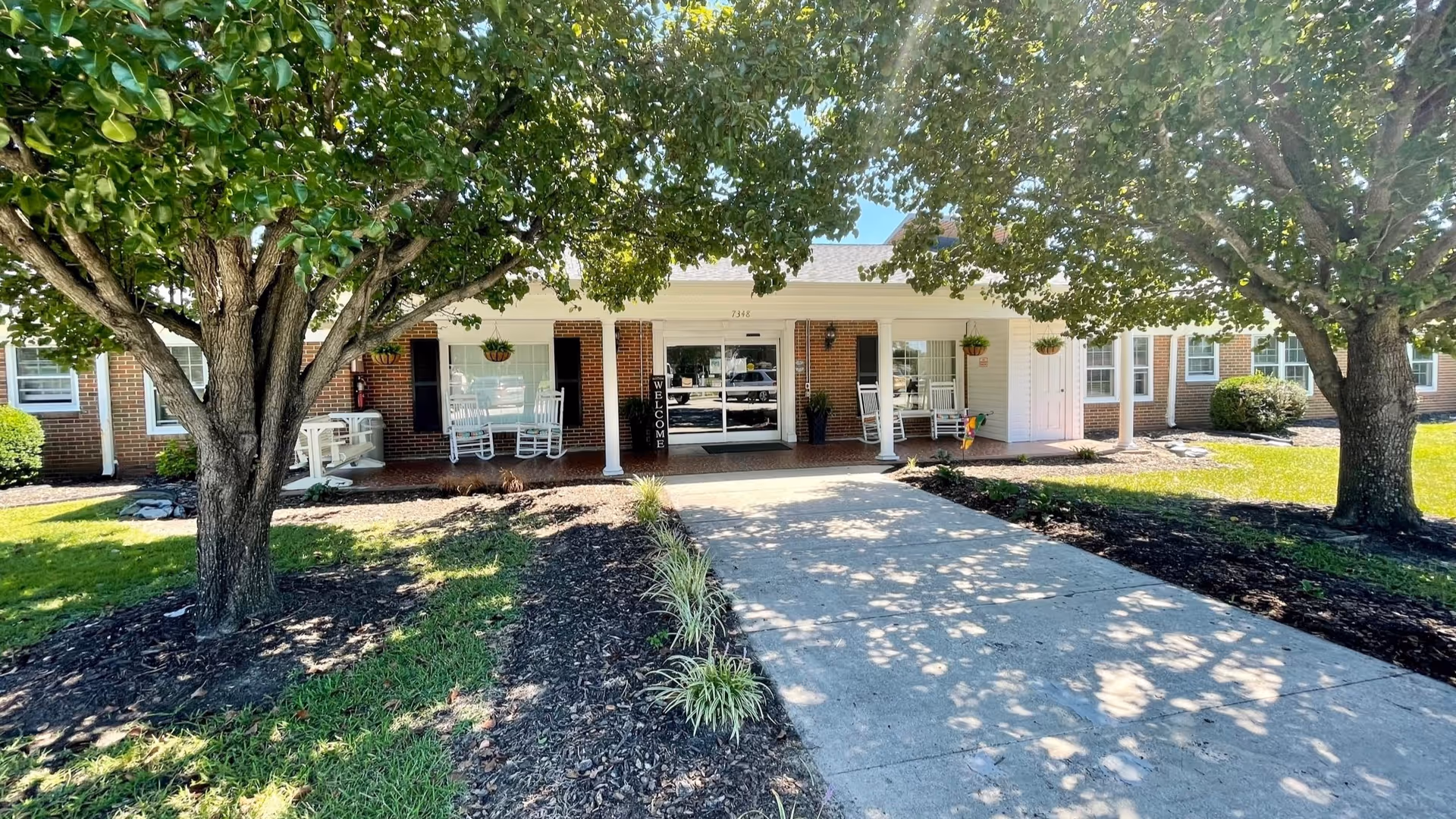 Front entrance of a nursing home with a covered porch, rocking chairs, a 'Welcome' sign, and a tree-lined walkway.