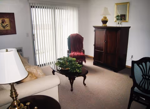 A living room with a beige sofa, a wooden coffee table with a green plant, a wooden cabinet with a lamp and a mirror on top, a chair with a red throw draped over it, vertical blinds covering a sliding glass door, and a framed floral picture on the wall.