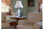 A cozy living room area with two patterned armchairs facing each other, a wooden side table between them holding a white decorative lamp, a ceramic dish, and a small decorative item. The walls are painted green, and there are two windows with beige curtains allowing natural light to enter. A framed painting hangs on the wall above one of the chairs.