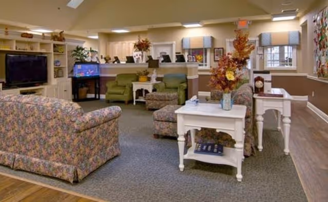 A cozy living room area in a senior living facility with floral patterned armchairs and sofas, a white side table with a vase of flowers, a large flat-screen TV on a white entertainment unit, and a fish tank. The room has beige walls with white trim, carpeted flooring, and windows with blue valances.