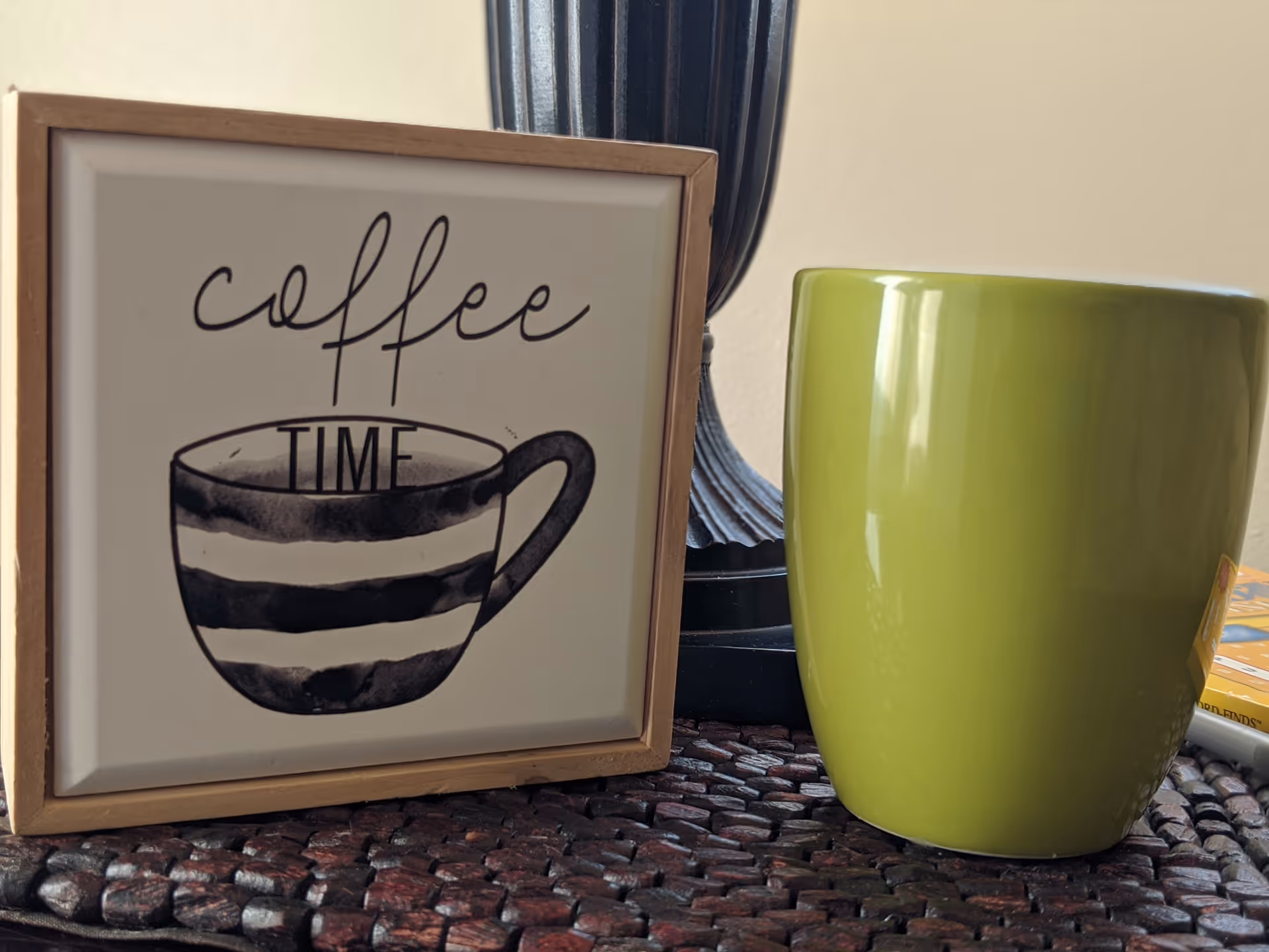 A framed decorative sign with a black and white striped coffee cup illustration and the words 'coffee TIME' written above it, placed next to a large green coffee mug on a woven surface.