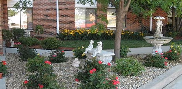 Landscaped front garden with rose bushes, stone benches, a fountain, and a brick building facade.