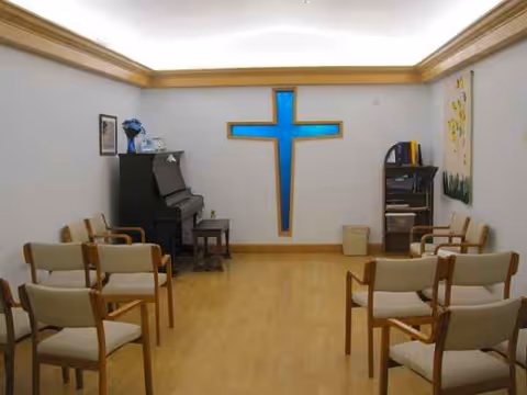 Chapel-like room with a blue cross on the wall, pianos, and rows of chairs.