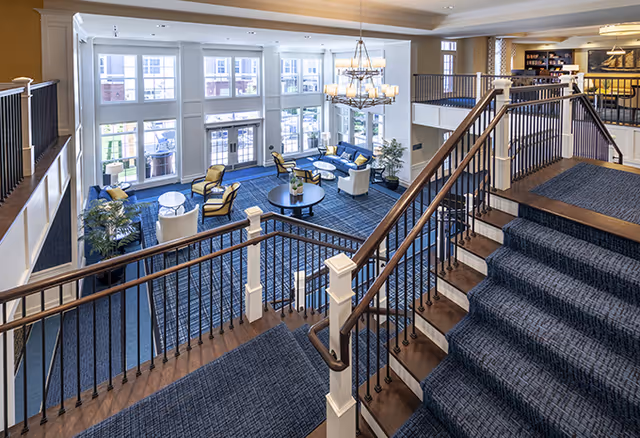 Interior view of a senior living facility showing a spacious common area with large windows letting in natural light. The area features blue carpeted floors, comfortable seating including armchairs and sofas arranged around round tables, and a decorative chandelier hanging from the ceiling. A staircase with wooden handrails and blue carpeted steps leads down to the seating area. There are plants and bookshelves in the background.