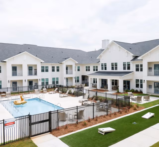 Outdoor courtyard area of a senior living facility featuring a fenced swimming pool with a golden inflatable duck, lounge chairs, a covered patio with seating, and a grassy area with cornhole boards. The building surrounding the courtyard is two stories with white siding and multiple windows and balconies.