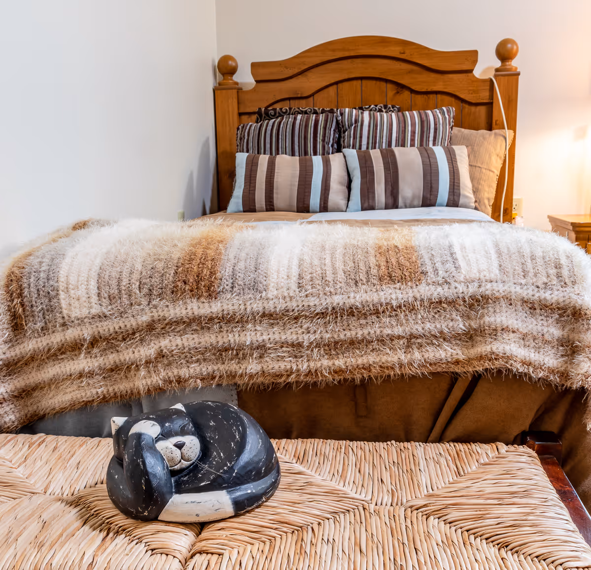 A cozy bedroom with a wooden headboard, striped pillows, a fuzzy beige-and-brown throw on the bed, and a woven bench topped by a small curled cat sculpture.