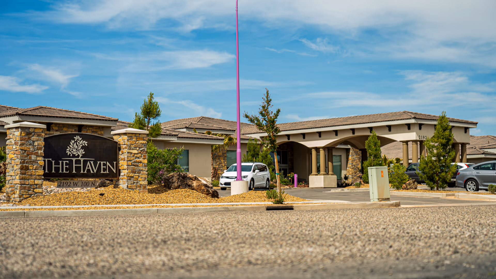 Front entrance of The Haven senior living community with a stone sign, covered porte-cochère, parked cars, and a clear blue sky.