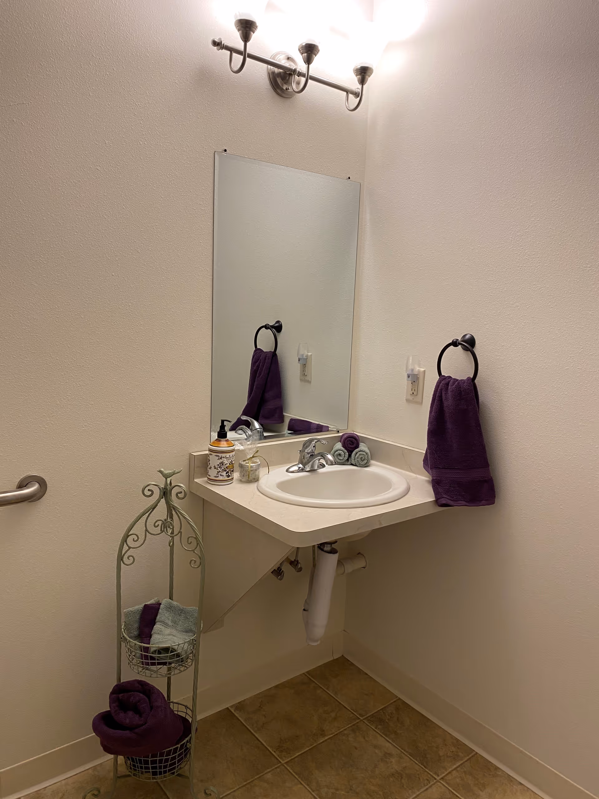 A small bathroom corner with a wall-mounted sink and a large rectangular mirror above it. There is a silver faucet, a soap dispenser, and a small container on the sink. A purple hand towel hangs on a black ring holder attached to the wall. Next to the sink is a decorative metal stand holding rolled towels in purple and light green. The floor is tiled, and the walls are painted white. A three-bulb light fixture is mounted above the mirror.
