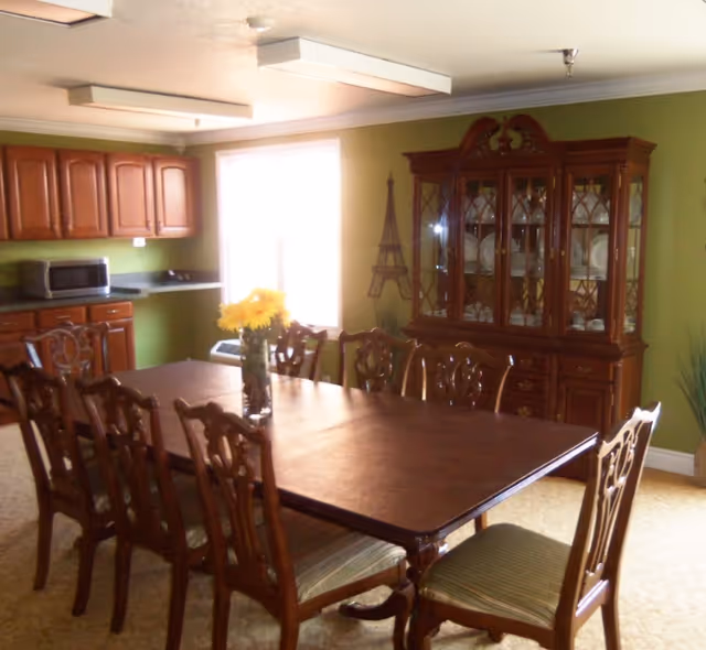 A dining room with a long wooden table surrounded by eight wooden chairs with cushioned seats. A vase with yellow flowers is placed in the center of the table. In the background, there is a wooden china cabinet displaying plates and glassware, and a kitchen area with wooden cabinets, a microwave, and a countertop. The walls are painted green and there is a window letting in natural light.
