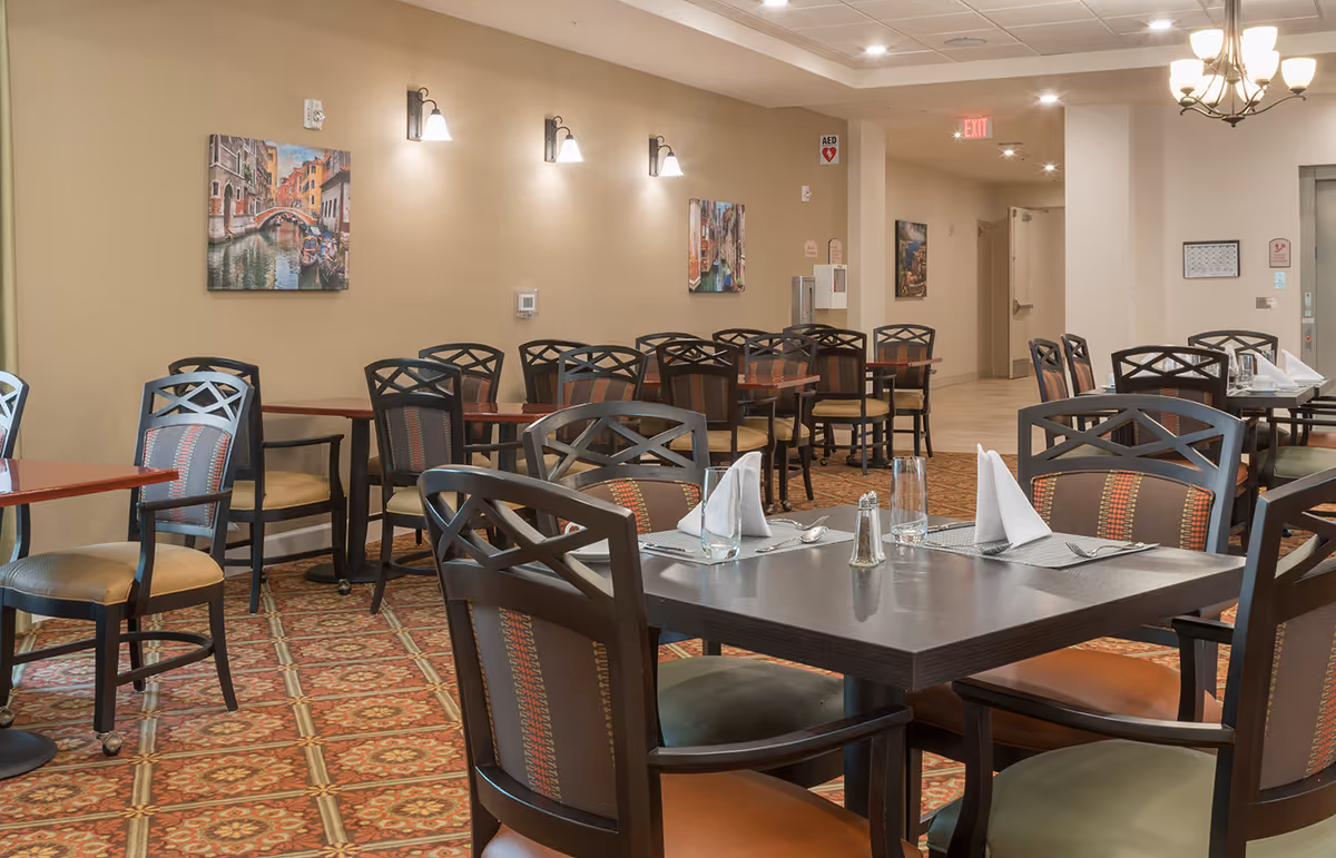 A dining room with multiple tables and chairs arranged neatly. The tables are set with placemats, napkins, glasses, and silverware. The room has beige walls adorned with paintings and wall-mounted lights. The floor is covered with a patterned carpet, and there is an exit sign visible in the background.