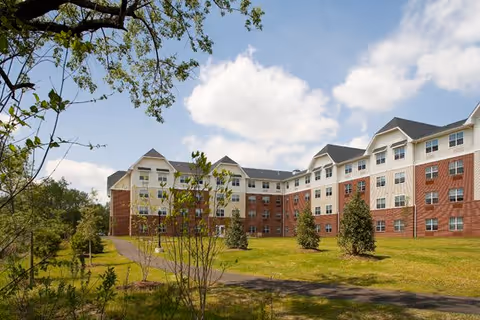Exterior view of a multi-story senior living facility building with a combination of red brick and light-colored siding. The building is surrounded by a grassy area with small trees and a paved walkway. The sky is partly cloudy.