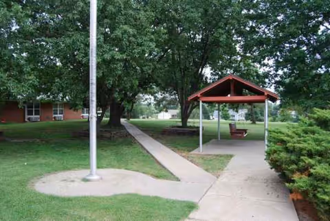 A shaded outdoor courtyard with a gazebo, bench, walkway, and trees beside a brick building.