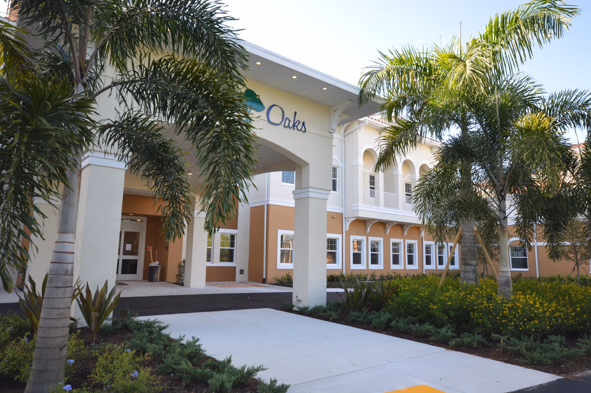 Exterior view of the Grand Oaks of Palm City senior living facility showing the entrance with an archway, palm trees, and landscaped greenery under a clear sky.
