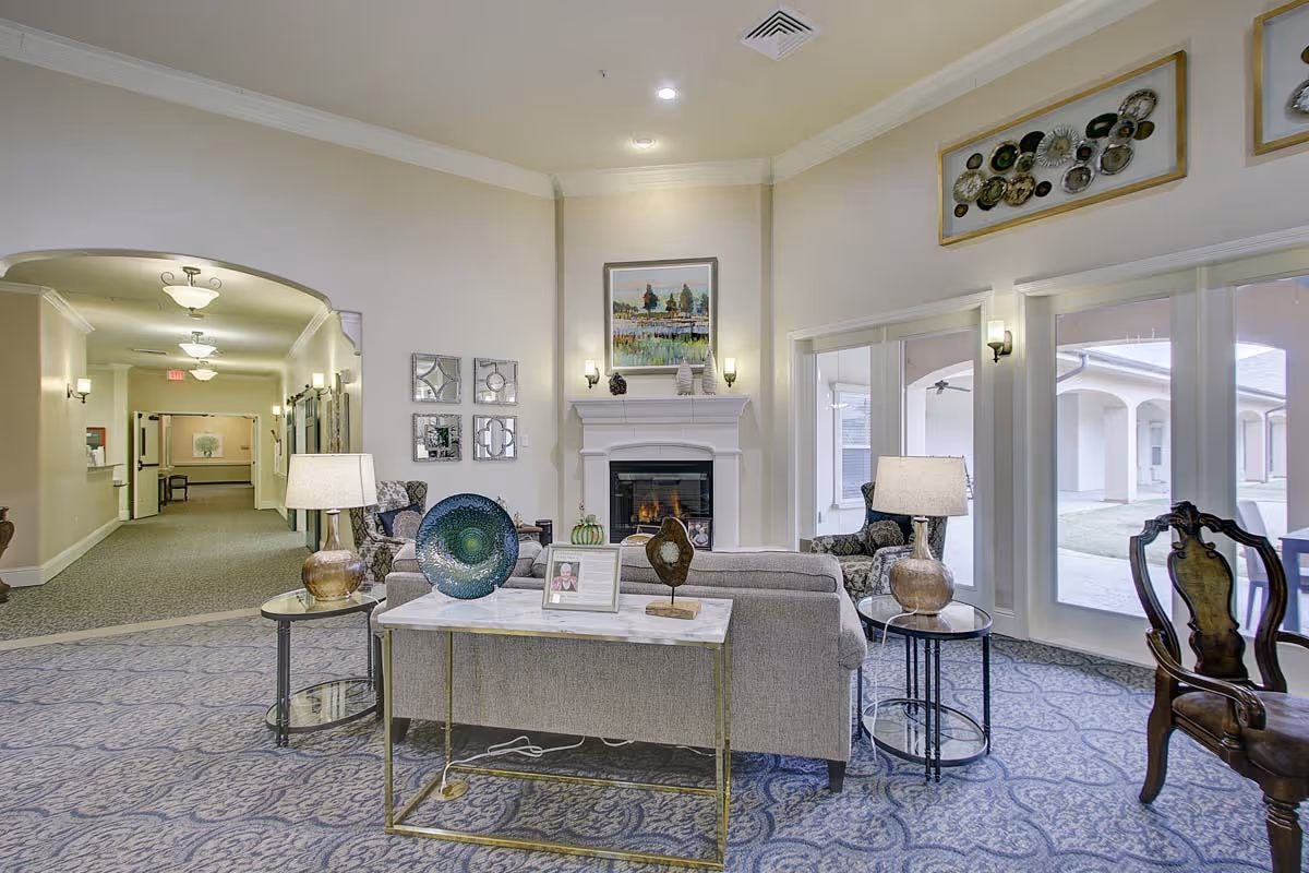 A cozy living room area in The Oaks At Flower Mound featuring a gray sofa, two patterned armchairs, a white fireplace with a painting above it, decorative plates on the wall, two table lamps on side tables, and large windows showing an outdoor courtyard.