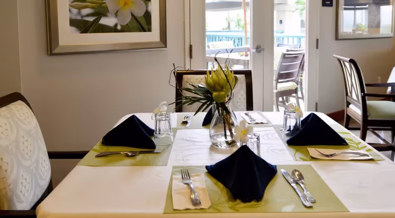 A dining table set for four with green placemats, navy folded napkins, silverware and a vase of flowers in a bright dining room with chairs and a door to an outdoor patio.