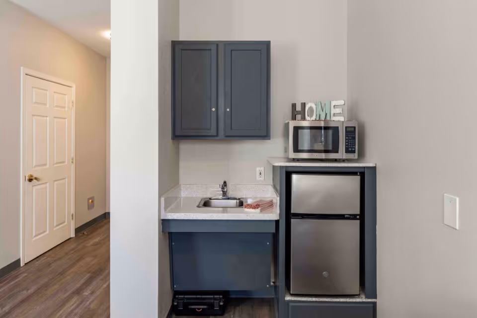 A small kitchenette area with a stainless steel mini refrigerator and microwave on top. Above the sink is a dark gray cabinet, and the countertop has a small sink with a faucet. The word HOME is displayed on top of the microwave. The floor is wood, and there is a white door visible in the background.