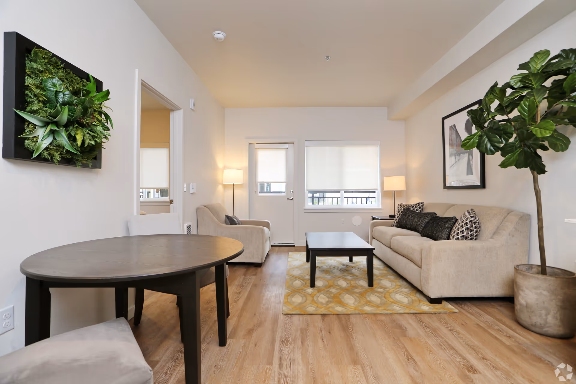 A modern living room with light beige walls and wood flooring. The room features a beige sofa with black and patterned cushions, a matching armchair, a black coffee table on a yellow patterned rug, and two floor lamps. There is a small round dining table with chairs in the foreground, a large potted plant on the right, and a framed picture on the wall. A door and window with white blinds are visible at the back.