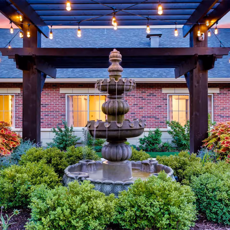 A tiered stone fountain surrounded by shrubs under a wooden pergola with string lights in front of a brick building.