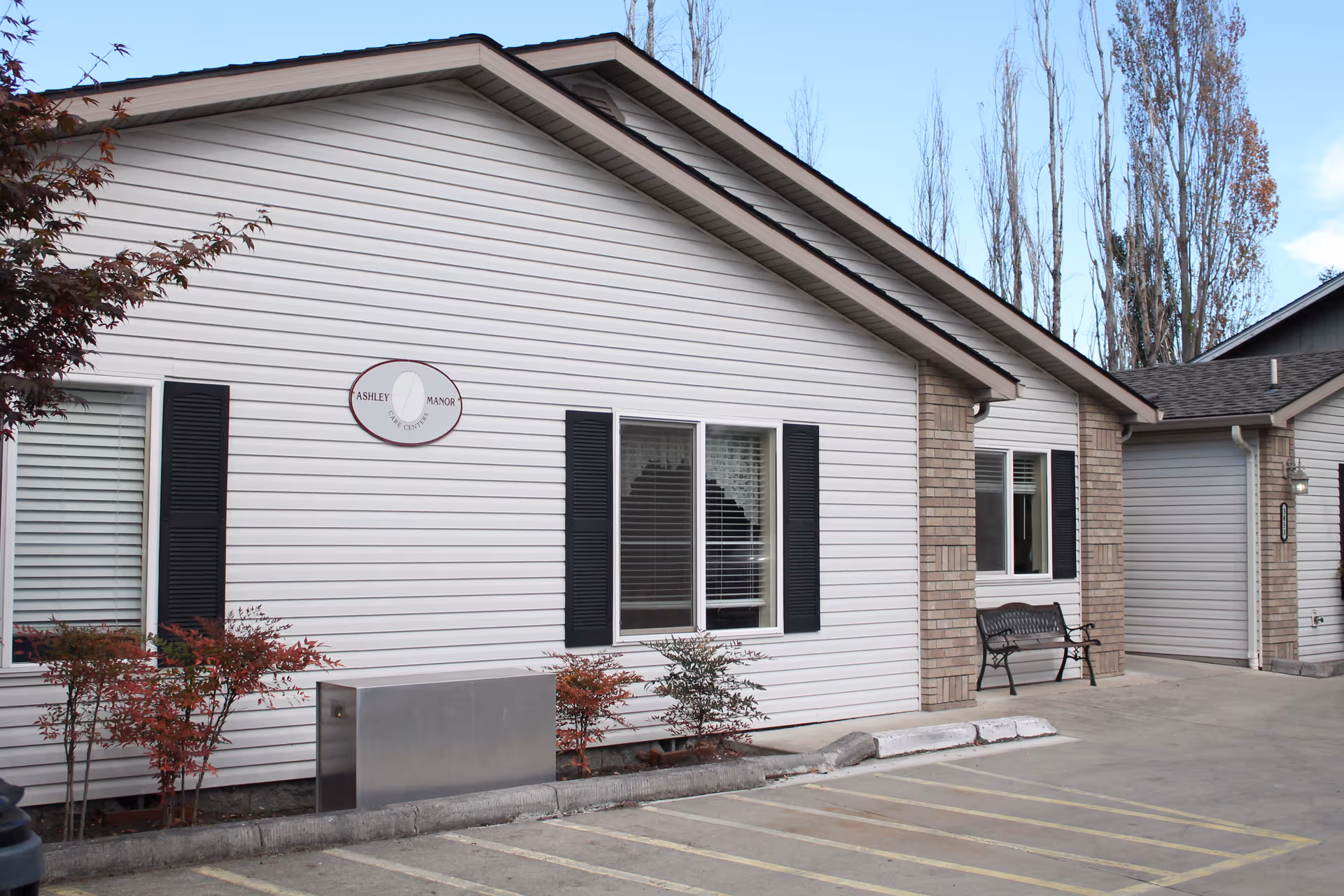 Front of a single-story white siding building with windows, black shutters, a bench, small shrubs, and a parking area.