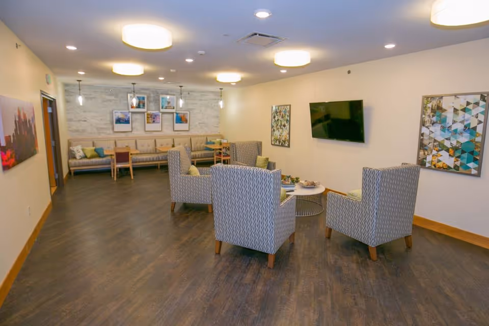 A well-lit common area with four patterned armchairs arranged around a small round coffee table. The room has wood flooring, a wall-mounted flat screen TV, and framed artwork on the walls. In the background, there is a long cushioned bench with several chairs and tables, pendant lights hanging from the ceiling, and additional framed pictures on the wall.