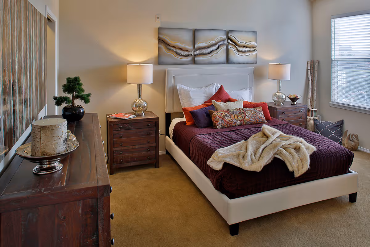 A cozy bedroom with a white upholstered bed frame and a dark purple quilted bedspread. The bed is adorned with multiple colorful pillows and a beige throw blanket. On either side of the bed are wooden nightstands with matching lamps. A large window with blinds lets in natural light. A wooden dresser with decorative items, including a small potted plant and a candle, is visible on the left side of the room. Above the bed hangs a three-piece abstract artwork.