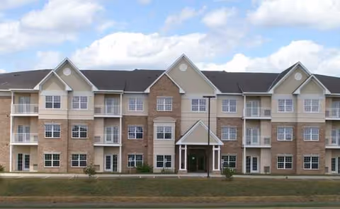Three-story senior living building with beige siding, balconies, and a central entrance.