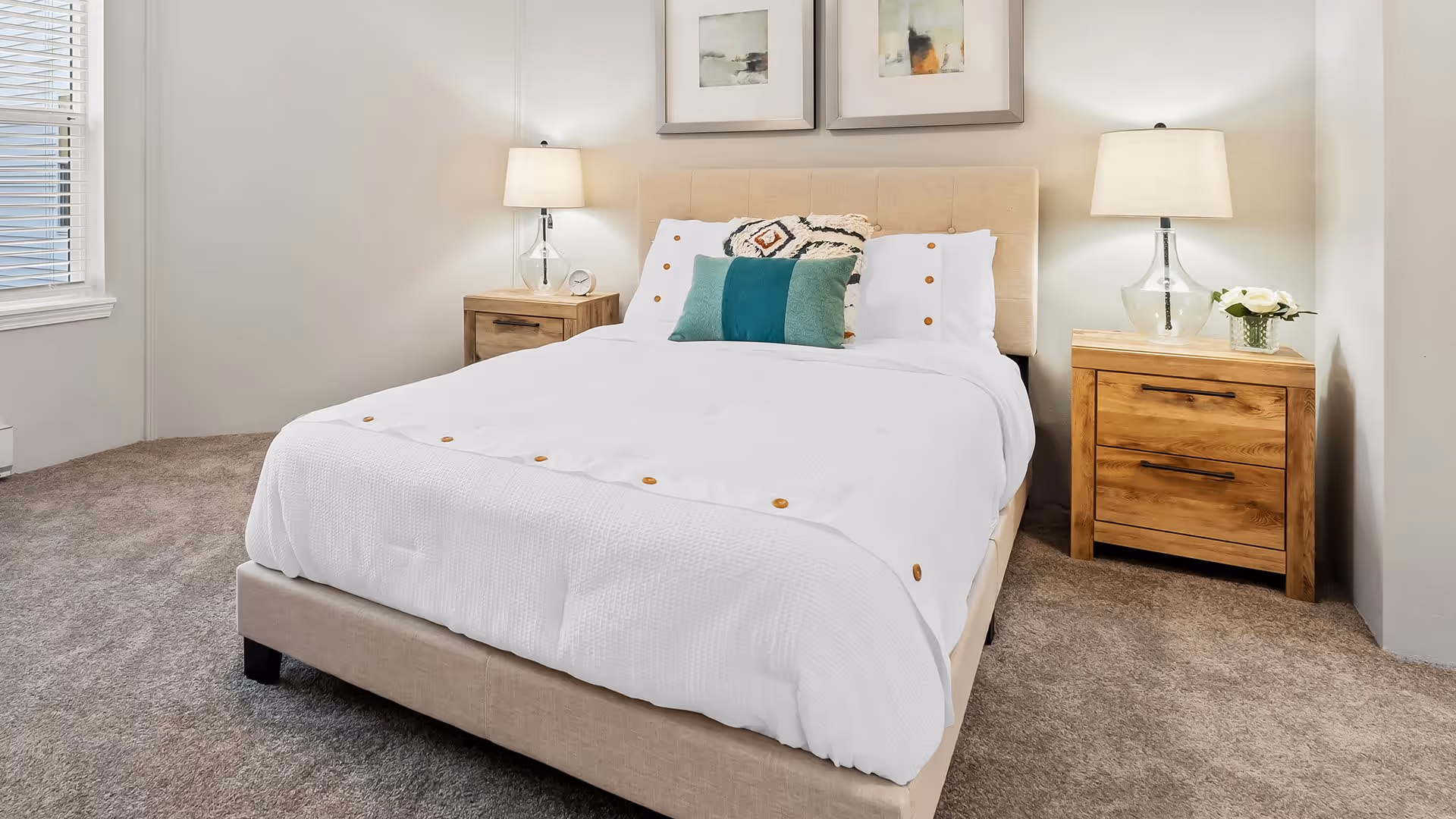 A neatly made bed with white bedding and decorative pillows in a bedroom. The bed has a beige upholstered headboard and is flanked by two wooden nightstands, each with a lamp and small decorative items. The room has beige carpet and light-colored walls with two framed pictures hanging above the bed.