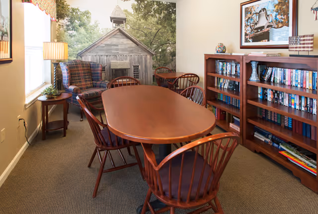 A cozy interior room with a wooden oval table surrounded by six wooden chairs with dark cushions. To the left, there is a plaid upholstered armchair next to a small round side table with a lamp and a plant. The back wall features a large mural of a rustic wooden building with trees. On the right side, there are two wooden bookshelves filled with books and DVDs, with a framed picture and small decorative items on top.