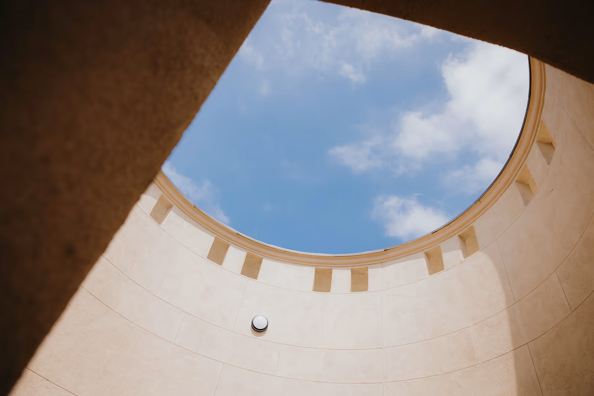 View up through a circular atrium oculus showing blue sky above curved beige walls.