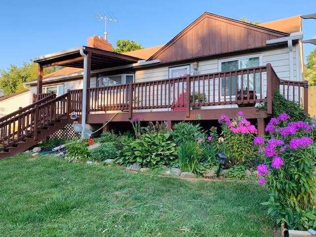 Exterior view of a single-story house with a brown wooden deck and railing. The house has a sloped roof and several windows. In front of the deck, there is a garden with green plants and vibrant purple flowers. The sky is clear and blue.
