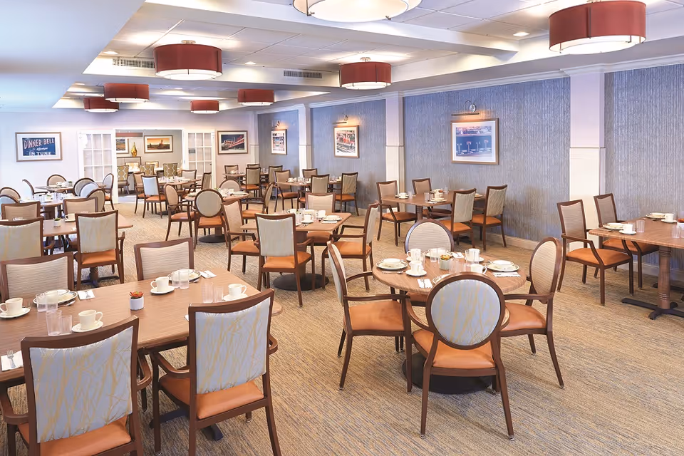 A spacious dining room with multiple wooden tables and chairs arranged neatly. Each table is set with white cups, plates, and utensils. The room features soft lighting with round ceiling fixtures and framed pictures on the walls. The carpet is beige with a subtle pattern, and the overall ambiance is warm and inviting.