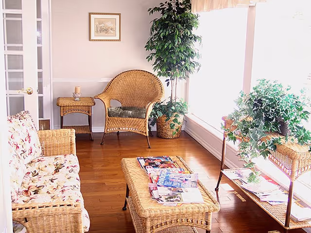 A bright living room area with wicker furniture including a floral cushioned sofa, a wicker chair with a cushion, a wicker coffee table with magazines on it, and a wicker side table with a candle. There are several green plants near a large window letting in natural light, and a framed picture hangs on the wall.