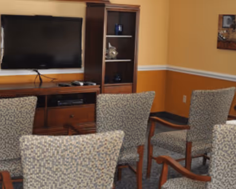Upholstered chairs arranged facing a wall-mounted TV and wooden entertainment cabinet in a common room.