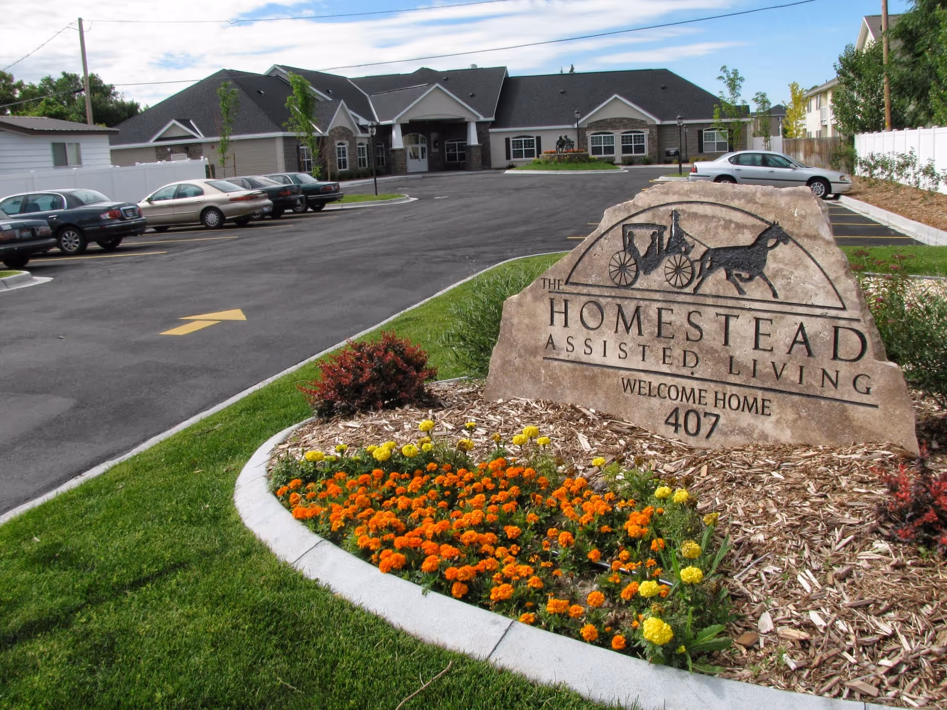 Stone entrance sign for 'Homestead Assisted Living' beside a flower bed with the facility building and parking lot visible behind it.