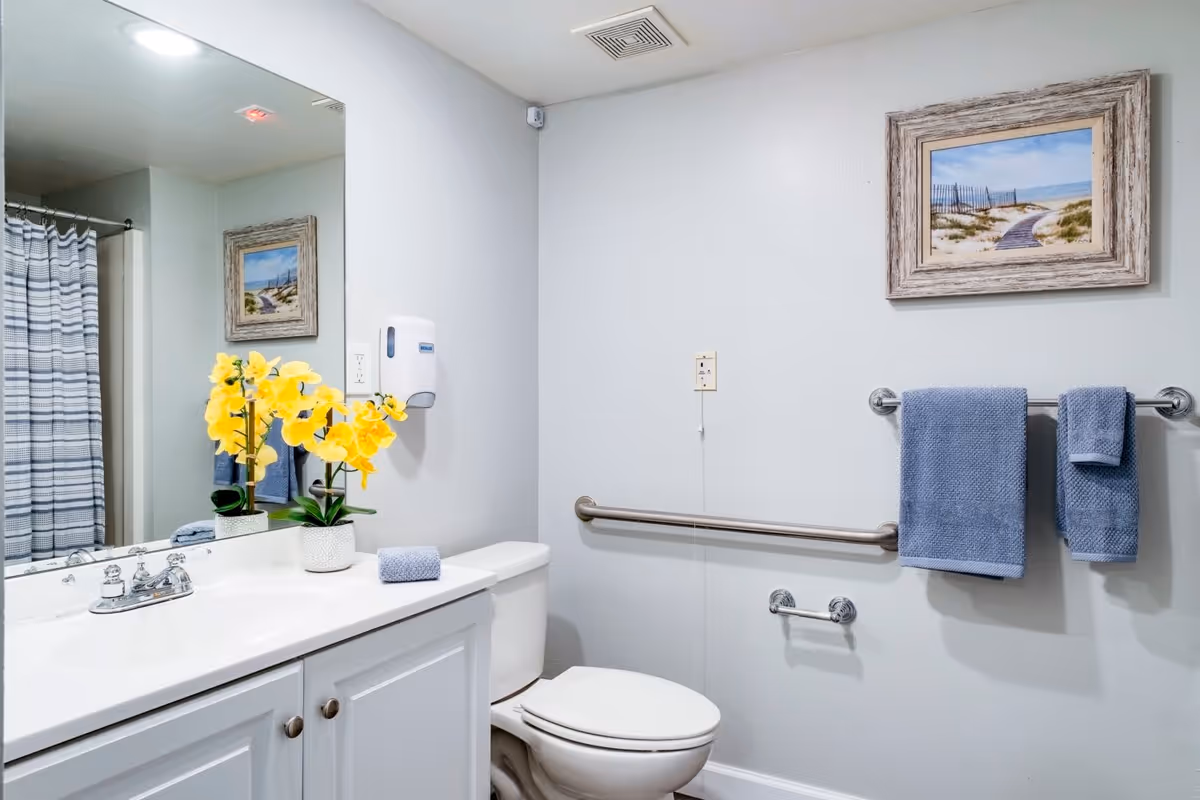 A clean and bright bathroom with a white vanity and sink, a toilet, and a shower with a striped curtain. There is a large mirror above the sink reflecting a framed beach-themed picture on the opposite wall. Yellow flowers in a white pot and a folded blue towel are placed on the vanity. Blue towels hang on a towel rack next to the toilet, and a grab bar is installed on the wall for support.
